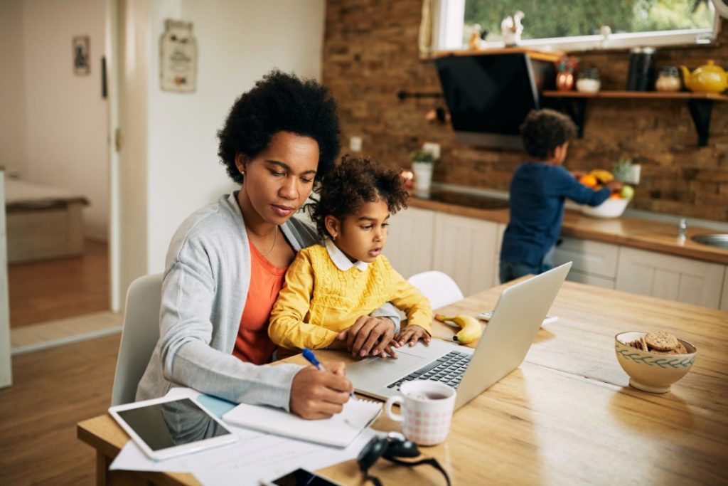 Woman writing notes while holding a young child at the kitchen table